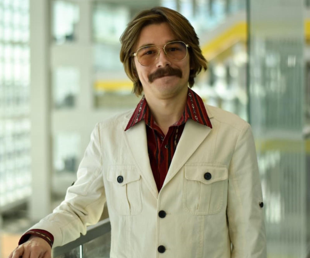 Maurycy Krzyżanowski, doktorant Uniwersytetu Michigan, w beżowej marynarce i koszuli we wzory, w okularach przeciwsłonecznych, z wąsami. Stoi w nowoczesnym, jasnym wnętrzu, opierając się o barierkę. Dowiedz się więcej o jego doktoracie w USA na Super Biznes.