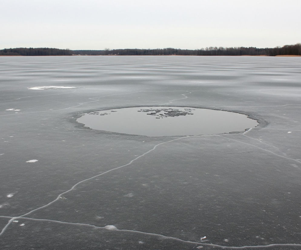 Zamarznięte jezioro w szarej tonacji, rozciąga się od pierwszego planu po horyzont. Na jego powierzchni widoczne są pęknięcia lodu i dwie okrągłe, ciemne plamy otwartej wody, z czego jedna, większa, znajduje się na pierwszym planie, a druga, mniejsza, dalej w tle. Brzegi jeziora otoczone są ciemnym lasem, a niebo nad nim jest jednolicie jasnoszare.