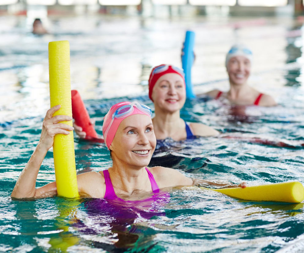 Trzy uśmiechnięte kobiety w czepkach i okularach pływackich, zanurzone w basenie, wykonują ćwiczenia z żółtymi i czerwonymi wałkami do aqua aerobiku. Wzmacniają swoje serca aktywnością fizyczną, o czym dowiesz się więcej na Poradnik Zdrowie.