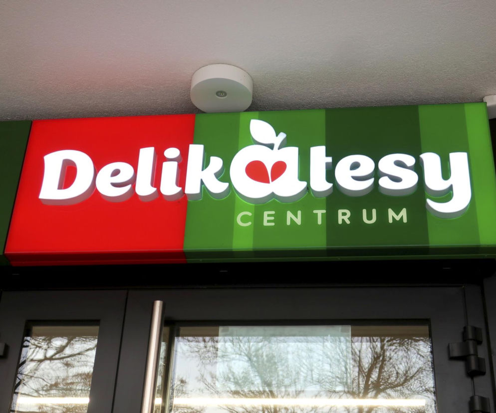 Szyld sklepu Delikatesy Centrum na czerwono-zielonym tle nad wejściem. Białe, podświetlane litery logo sieci handlowej Delikatesy Centrum, której restrukturyzacji i zamykania placówek poświęcony jest artykuł, dostępny na portalu Super Biznes.