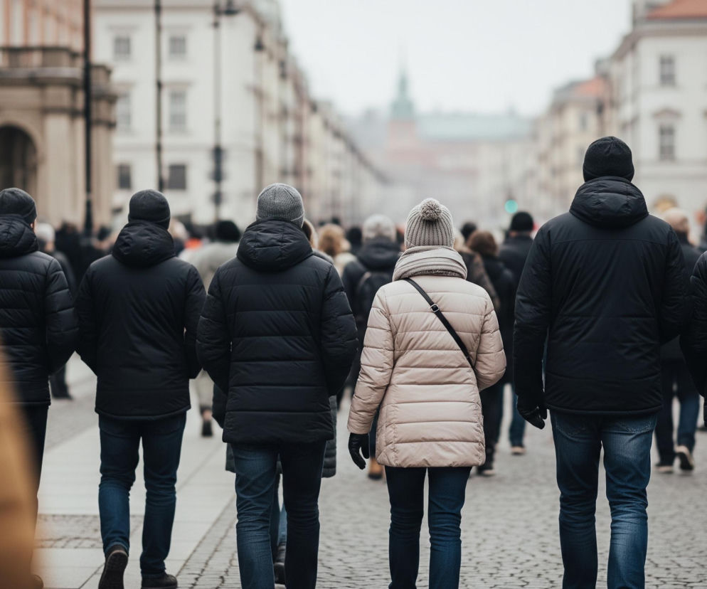 Grupa osób idzie ulicą w pochmurny dzień, w kadrze widoczne są sylwetki od tyłu lub lekko z boku. Na pierwszym planie po lewej stronie znajduje się rozmazany fragment osoby w jasnobrązowej kurtce, natomiast w centrum i po prawej stronie widoczne są osoby w ciemnych kurtkach zimowych i czapkach, niektóre w dżinsach. Pośrodku grupy wyróżnia się kobieta w beżowej, pikowanej kurtce i szarej czapce z pomponem, z czarną torebką przewieszoną przez ramię. W tle widać kamienice o jasnych elewacjach i elementy miejskiej infrastruktury, jak latarnie, a na horyzoncie majaczy wieża.