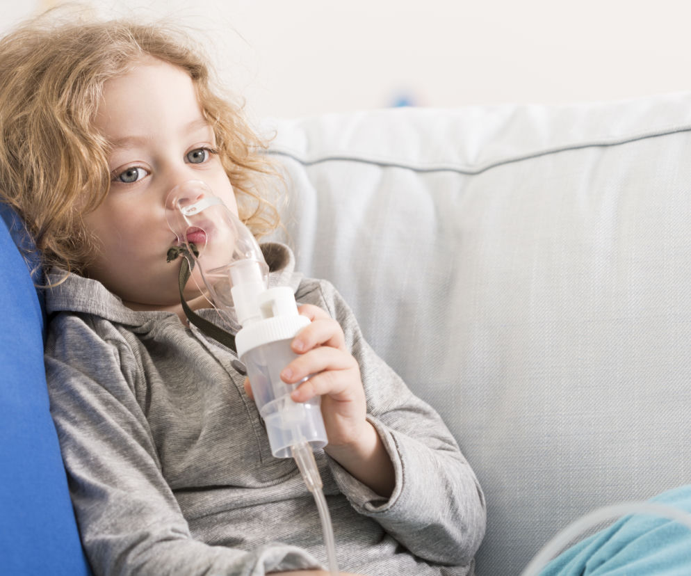 Dziecko z maseczką do inhalacji siedzi na kanapie. O organizacji domu w czasie choroby przeczytasz na SE.
