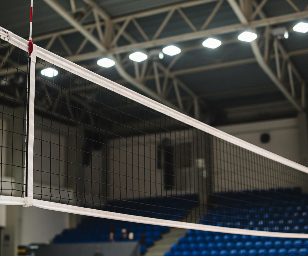 Czarna siatka do siatkówki, rozciągnięta wzdłuż. Górna krawędź siatki jest obszyta szeroką, białą taśmą, a po lewej stronie siatki widoczny jest pionowy biało-czerwony pręt, służący jako antena. Siatka jest przymocowana białymi linkami do słupków poza kadrem, a w tle widać ciemne trybuny z niebieskimi siedzeniami oraz konstrukcję dachu hali sportowej z jasnymi lampami.