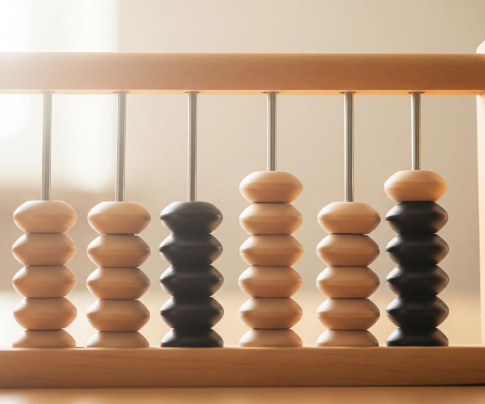 Drewniane liczydło z metalowymi prętami, na których znajdują się koraliki, wypełnia centralną część kadru. Rama liczydła ma jasnobrązowy kolor naturalnego drewna, a w jej górnej i dolnej części umieszczono pięć pionowych metalowych prętów. Na każdym pręcie zawieszonych jest pięć koralików w kształcie spłaszczonych owali; koraliki na drugim i piątym pręcie od lewej są czarne, natomiast pozostałe są jasnobrązowe. Całe liczydło spoczywa na drewnianej, beżowej powierzchni, a tło jest rozmyte, jasne i jednolite, z lekkim gradientem od jaśniejszego po lewej stronie do ciemniejszego po prawej.