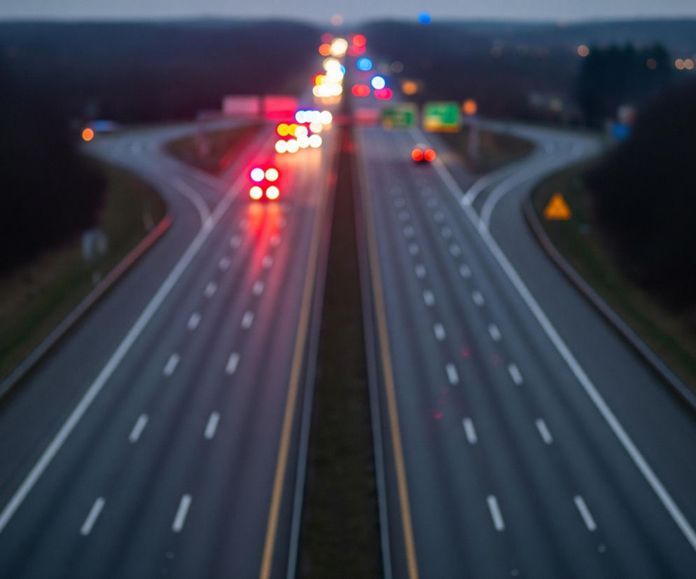 Asfaltowa autostrada z kilkoma pasami ruchu biegnie przez środek obrazu, rozwidlając się w tle. Na drodze widoczne są rozmyte, jasne światła samochodów, głównie czerwone tylne światła, oraz kilka białych i niebieskich świateł z przodu. Otaczające autostradę tereny pokryte są ciemnymi drzewami i trawą, a niebo nad horyzontem jest szare.