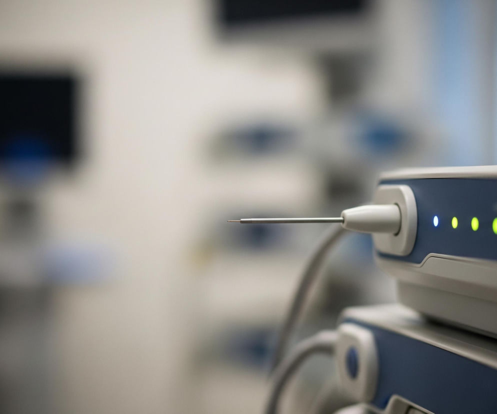 W centralnej części kadru znajduje się zbliżenie na część sprzętu medycznego, prawdopodobnie endoskopu, z ostrą metalową końcówką wystającą z białej obudowy, połączonej z niebiesko-białym urządzeniem. Na górnej, niebieskiej powierzchni urządzenia widoczne są cztery świecące diody: pierwsza od lewej jest jasnoniebieska, a trzy kolejne są jasnozielone. Urządzenie jest podłączone kablami, które częściowo widać w tle. Tło jest rozmyte, dominują w nim jasne, neutralne odcienie bieli i szarości, z kilkoma niewyraźnymi ciemniejszymi plamami, przypominającymi ekrany monitorów lub inne urządzenia, oraz niebieskimi elementami.