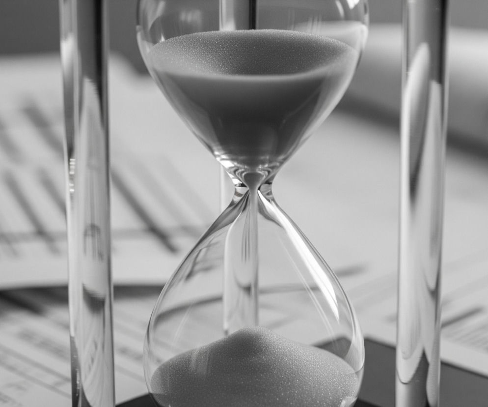 Na zdjęciu widoczna jest klepsydra, umieszczona centralnie, z widocznym przepływającym piaskiem. Jej szklane bańki są wypełnione piaskiem w górnej części, podczas gdy strumień piasku przesypuje się przez wąskie gardło do dolnej bańki, tworząc mały kopczyk. Obraz utrzymany jest w monochromatycznej palecie szarości, a w tle widać rozmyte, zadrukowane kartki papieru, które sugerują kontekst biurowy lub pracy.
