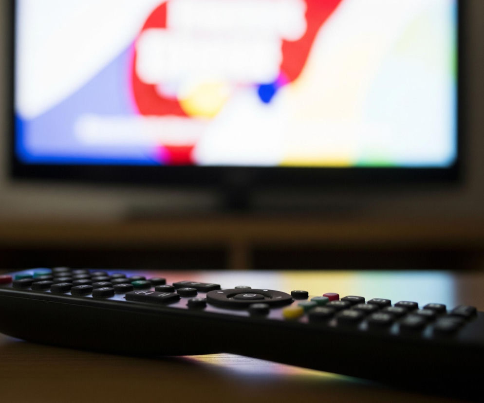 Czarny pilot do telewizora leży na jasnobrązowej, drewnianej powierzchni, znajdującej się na pierwszym planie obrazu. W tle, rozmyty ekran telewizora wyświetla abstrakcyjną kompozycję barwnych kształtów w odcieniach czerwieni, błękitu, żółci i bieli. Przedmiotem centralnym jest pilot, który skierowany jest lekko w prawo, ukazując rząd czarnych przycisków, a także kilka kolorowych, między innymi czerwony, żółty i zielony.