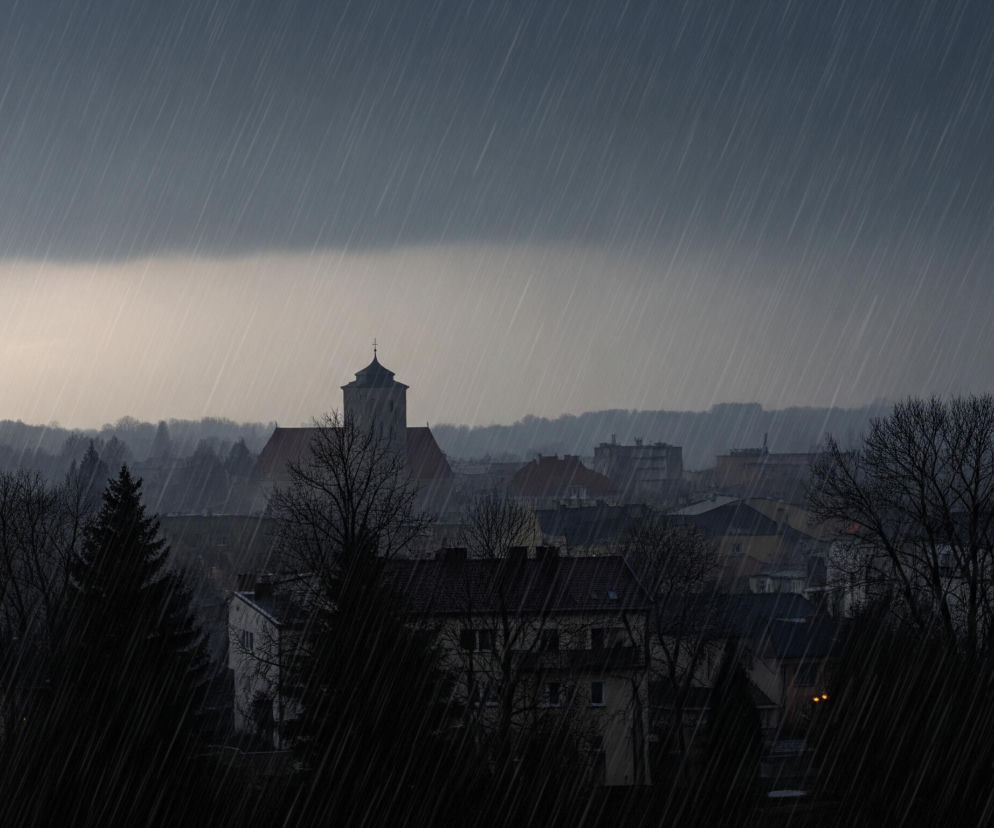 Panorama miejska podczas ulewnego deszczu, z dominującym szarym, zachmurzonym niebem. Na pierwszym planie widoczne są ciemne, bezlistne drzewa i iglaki, częściowo zasłaniające zabudowania mieszkalne. W tle, ponad dachami budynków, wznosi się ciemny kościół z charakterystyczną wieżą zakończoną spiczastym dachem i krzyżem. Cała scena jest zalana smugami deszczu, nadając jej ponury i melancholijny nastrój.