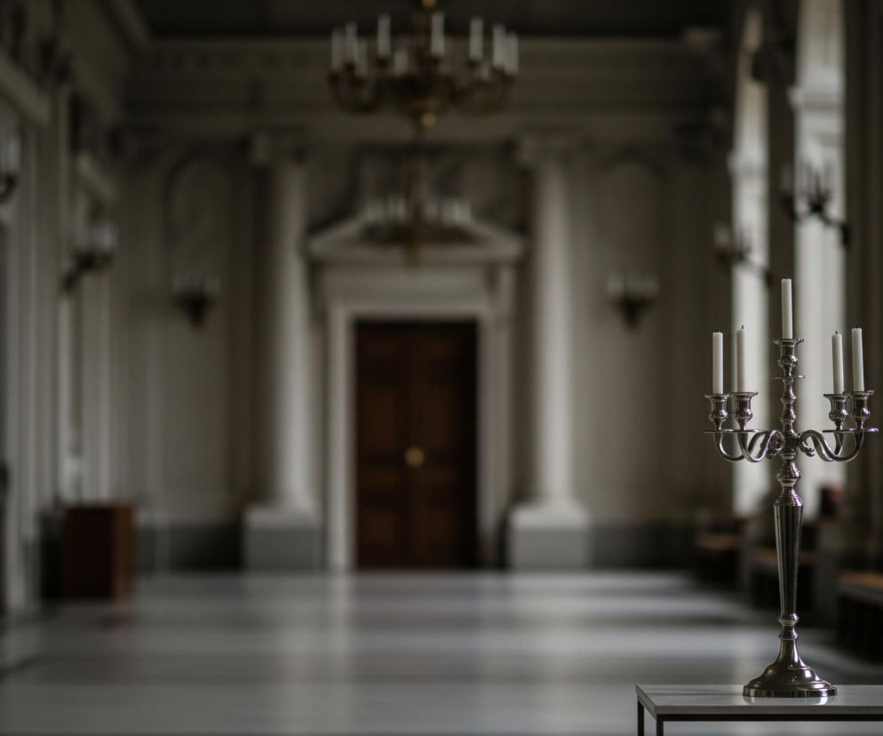 Na pierwszym planie, z prawej strony, stoi srebrny, lśniący świecznik z pięcioma białymi, niezapalanymi świecami, umieszczony na prostokątnym, białym stole. Tło stanowi długi, przestronny korytarz lub sala o wysokim suficie i jasnych ścianach, z rzędami kolumn i rzeźbionymi elementami architektonicznymi. Na ścianach widać ciemne kinkiety, a na suficie wiszą rozmazane żyrandole, wszystkie w ciemnych odcieniach, zlewające się z otoczeniem. Na końcu korytarza znajduje się wysokie, ciemne, dwuskrzydłowe drzwi, otoczone białymi kolumnami.