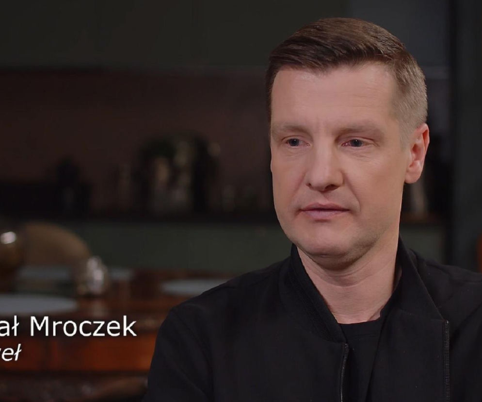 Portret Rafała Mroczka, aktora serialu M jak miłość, ubranego w czarną kurtkę, patrzy w prawo z lekko zamyślonym wyrazem twarzy. Na tle rozmytego wnętrza i napisu Rafał Mroczek Paweł. O jego szczerych wypowiedziach o serialowej postaci przeczytasz na Super Seriale.