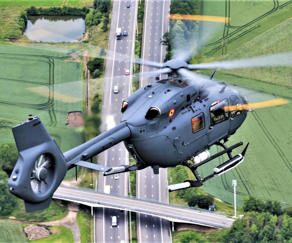 Wojskowy śmigłowiec Airbus H145M w locie nad zielonymi polami i autostradą, prezentujący belgijskie oznaczenia. Maszyna symbolizuje modernizację floty obronnej, o której więcej przeczytasz na naszym portalu.