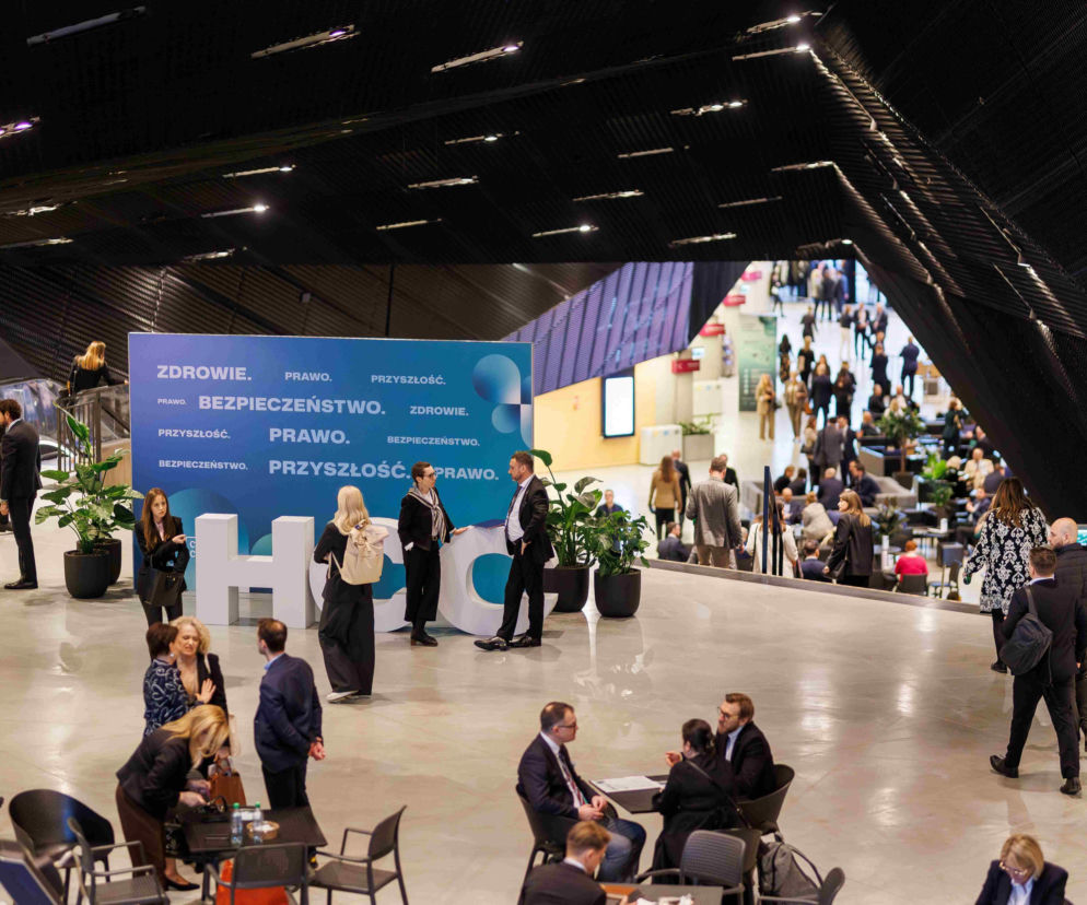 Uczestnicy XI Kongresu Wyzwań Zdrowotnych w Katowicach dyskutują w przestronnej hali. Na pierwszym planie widoczni są ludzie w swobodnych rozmowach przy stolikach, natomiast w tle znajduje się duży, niebieski baner z hasłami Zdrowie. Prawo. Przyszłość. Bezpieczeństwo.. Na portalu Poradnik Zdrowie znajdziesz więcej informacji o takich wydarzeniach.