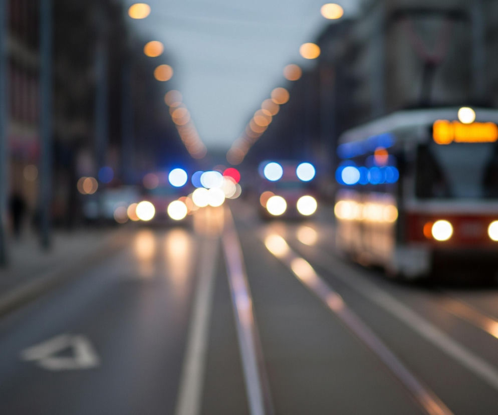 Rozmyte, nocne ujęcie miejskiej ulicy z widocznym w oddali tramwajem i samochodami. Na pierwszym planie po prawej stronie znajduje się rozmazany tramwaj, z wyraźnie widocznymi, jasnymi, żółto-pomarańczowymi światłami z przodu oraz rzędami niebieskich świateł po boku. Centralnie na drodze biegną metalowe szyny tramwajowe, rozjaśnione odbiciami świateł. W oddali, na ulicy, widać inne pojazdy, oświetlone rozmazanymi światłami o różnych kolorach, w tym czerwonym i niebieskim, a także pomarańczowym od latarni ulicznych, które tworzą świetliste kręgi. Po obu stronach ulicy znajdują się zamazane budynki i sylwetki przechodniów, wtapiające się w ciemne otoczenie.