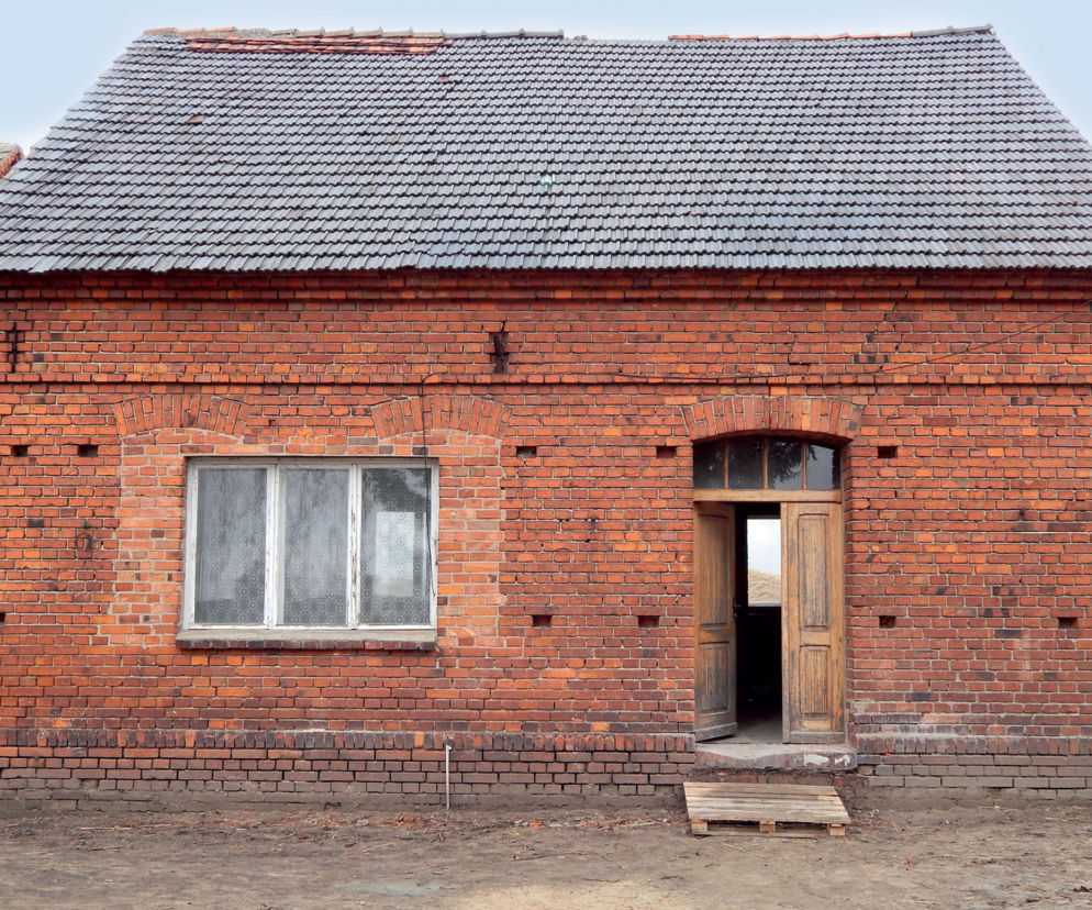 Stary, zrujnowany dom z czerwonej cegły i dachem z szarej dachówki ceramicznej, przed metamorfozą w piękne siedlisko. Widać zniszczone okno i otwarte, drewniane drzwi. Więcej o tej niezwykłej przemianie przeczytasz na naszym portalu.
