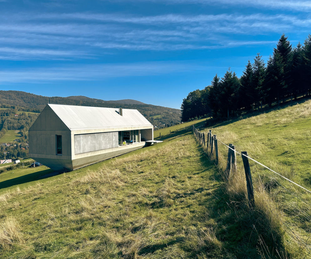 Arka Roberta Koniecznego w Brennej, dom z betonu i szkła uniesiony nad ziemią na zielonym zboczu Beskidu Śląskiego, na tle pasma gór i błękitnego nieba z delikatnymi chmurami. Więcej o tym wyjątkowym projekcie przeczytasz na Architektura Murator Plus.