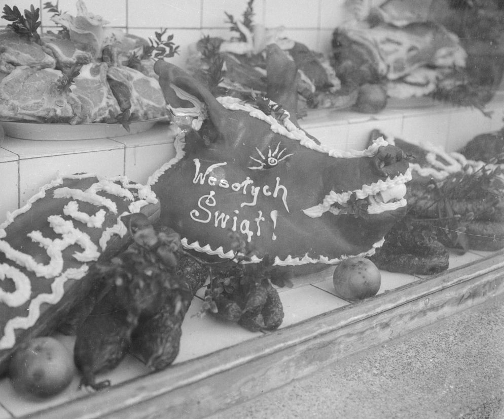 Czarno-białe zdjęcie prezentuje wystawę sklepu mięsnego w PRL z wyrobami na Wielkanoc. Widoczne są: głowa świni z napisem Wesołych Świąt!, wędliny oraz surowe mięso. Fotografia oddaje realia świąt w Polsce Ludowej, o których więcej przeczytasz na Super Biznes.