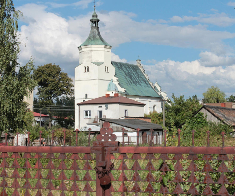 Biały kościół z zielonym dachem w Bolimowie widoczny zza metalowego płotu. O zabytkach miasta przeczytasz w Eska Łódź.