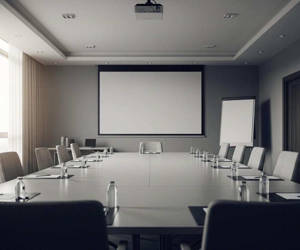 W centrum pomieszczenia znajduje się duży, prostokątny stół konferencyjny w jasnoszarym odcieniu, otoczony krzesłami z ciemniejszymi obiciami, tworząc układ w kształcie litery U. Na stole rozmieszczone są butelki z wodą, notesy i długopisy przed każdym miejscem. Wzdłuż lewej ściany, przez duże okno z jasnymi zasłonami, wpada rozproszone światło dzienne. Na przeciwległej ścianie widać duży, biały ekran projekcyjny z czarną ramą, a obok niego, po prawej stronie, stoi biała tablica suchościeralna na metalowym stojaku oraz ciemne drzwi. Na suficie rozmieszczone są liczne wpuszczane oprawy oświetleniowe, a także zamontowany projektor multimedialny.