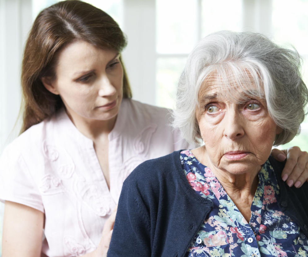 Starsza kobieta z siwymi włosami, wyraźnie zmartwiona lub przestraszona, obok której stoi młodsza kobieta w jasnej koszuli, delikatnie trzymająca ją za ramię. Zdjęcie symbolizuje wsparcie bliskich w obliczu demencji, o której więcej przeczytasz na Poradnik Zdrowie.