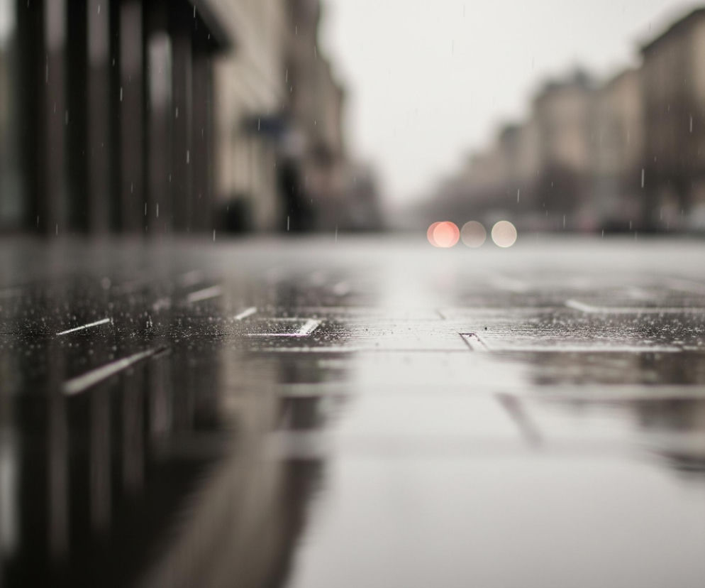 Mokry chodnik lub asfalt odbija światło, tworząc lśniące refleksy i zniekształcone odbicia budynków. Na pierwszym planie widać wyraźne, ciemne prostokątne płyty chodnika, pokryte licznymi kroplami deszczu. W tle, perspektywa ulicy rozciąga się w głąb, ukazując rozmyte kontury budynków po obu stronach. W oddali dostrzegalne są trzy rozmazane światła, w tym jedno wyraźnie czerwone i dwa jaśniejsze, prawdopodobnie pochodzące od pojazdów, a delikatne pionowe smugi świadczą o padającym deszczu.