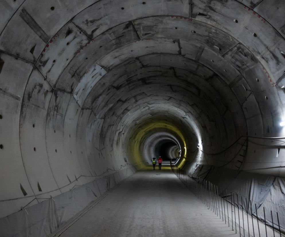 Wnętrze budowanego tunelu średnicowego w Łodzi, widoczni dwaj pracownicy w odblaskowych kamizelkach na tle betonowej, okrągłej konstrukcji. Wzdłuż ścian ułożone panele ochronne. To ważny element podziemnej architektury, o którym przeczytasz na Architektura Murator Plus.