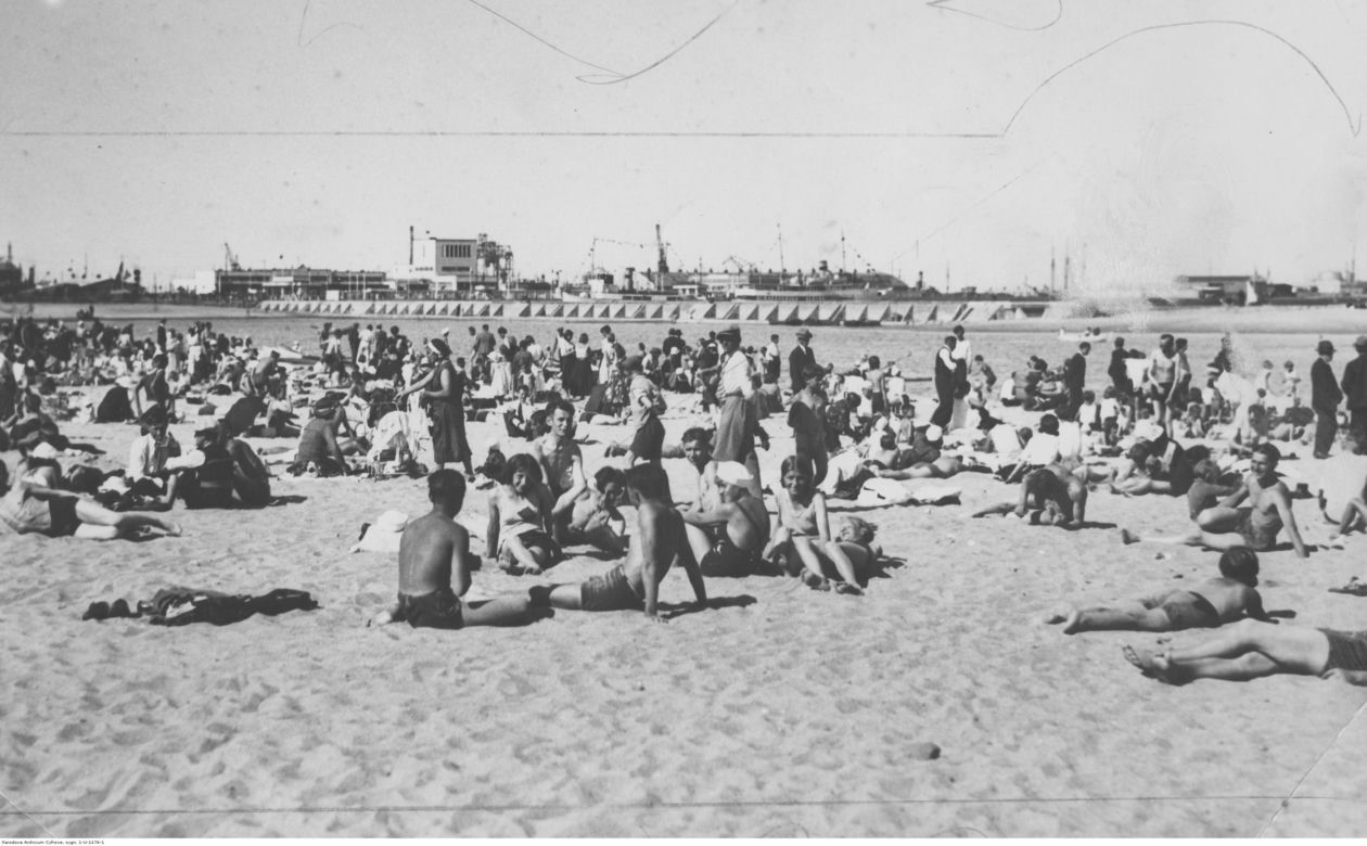 Plażowicze podczas wypoczynku nad morzem Bałtyckim. W tle port. Zdjęcie zrobiono w Gdyni pomiędzy 1923-1935 rokiem
