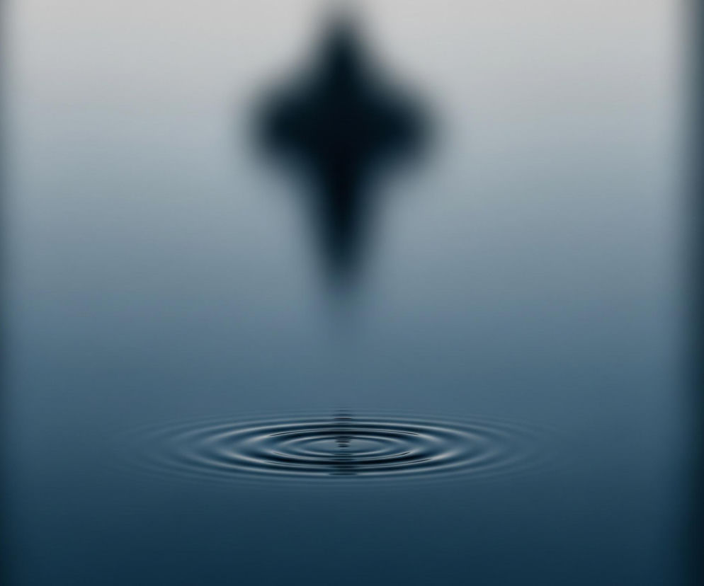 Centralnym elementem obrazu są koncentryczne fale na ciemnoniebieskiej powierzchni wody, rozchodzące się od małego punktu. Powyżej nich, w górnej części obrazu, widoczny jest pionowy, rozmyty, ciemny kształt przypominający grot strzały skierowany w dół, otoczony jaśniejszą, rozmytą poświatą. Obraz utrzymany jest w odcieniach niebieskiego i szarości, z ciemniejszymi partiami po bokach, przechodzącymi w jaśniejsze odcienie w centrum.