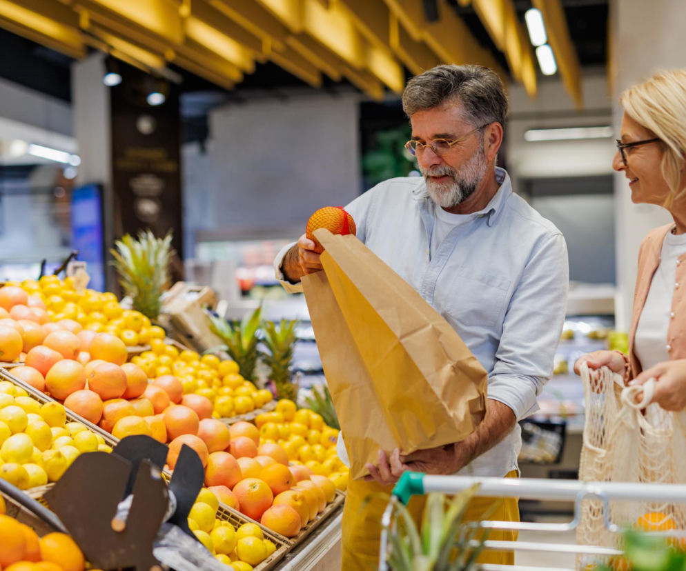 Starsza para w sklepie spożywczym, mężczyzna z siwą brodą i okularami wkłada grejpfruta do papierowej torby. Obok uśmiecha się kobieta w okularach. W tle mnóstwo owoców cytrusowych, takich jak pomarańcze, cytryny i grejpfruty. Poradnik Zdrowie radzi, jak wybierać produkty na zdrowe serce.