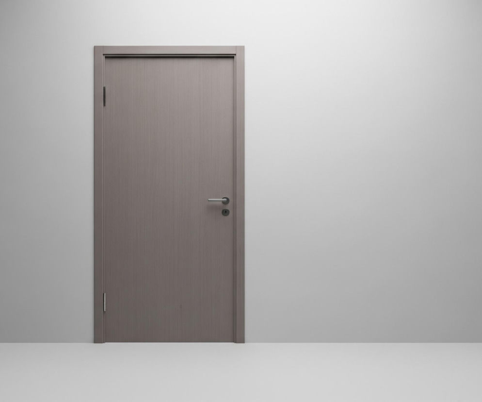 Pośrodku znajduje się zamknięte, szare skrzydło drzwiowe z delikatnym pionowym usłojeniem, zamontowane w ościeżnicy o identycznym kolorze i fakturze. Na drzwiach widoczne są srebrne, poziome klamka oraz okrągła, czarna rozeta na klucz, a także dwie ciemne zawiasy po lewej stronie. Drzwi są otoczone gładką, jasnoszarą ścianą, która rozciąga się również w prawą stronę, a u dołu przechodzi w białą podłogę. Lewa strona ściany jest nieco ciemniejsza i bardziej cieniowana niż prawa, która jest jasna i równomiernie oświetlona, co sugeruje źródło światła z prawej strony górnej części kadru.