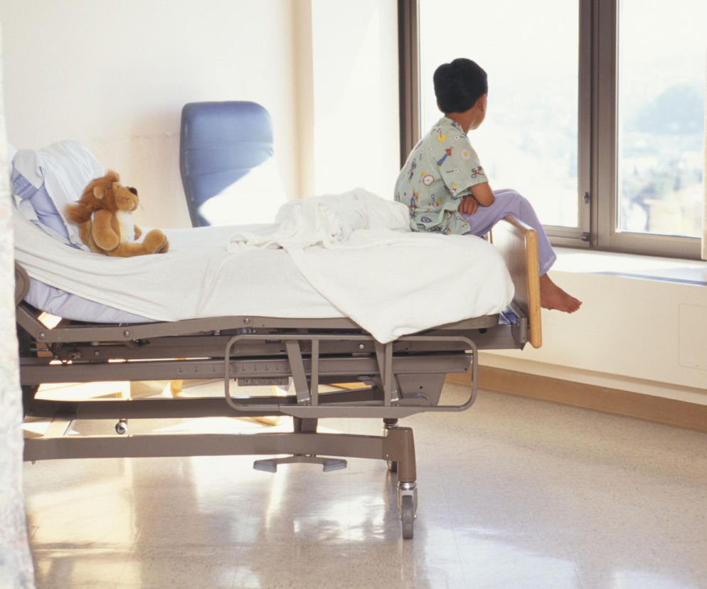 Chłopiec siedzący na łóżku szpitalnym tyłem do obiektywu, oparty o ramę łóżka, wpatrzony w okno, obok pluszowy lew. Artykuł na Poradnik Zdrowie omawia sildenafil jako nowe rozwiązanie w leczeniu chorób rzadkich u dzieci.