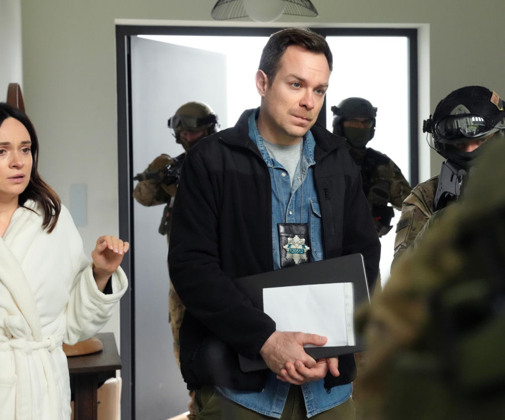 Funkcjonariusz policji w mundurze, z odznaką Policja 120512, stoi z teczką w dłoni, patrząc na zaskoczoną kobietę w białym szlafroku. Za nim widać umundurowanych policjantów z bronią, co zwiastuje kłopoty Rawiczów w serialu Barwy szczęścia. Więcej o tym na stronie Super Seriale.