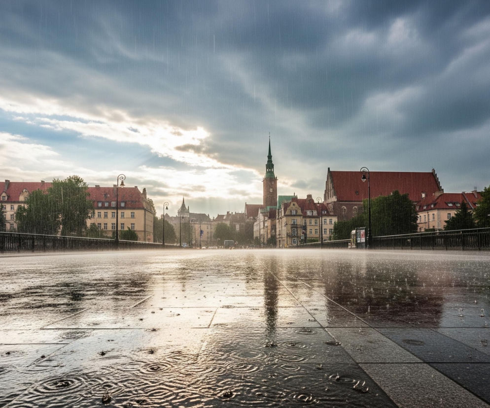 Szeroki plan przedstawia miejski krajobraz w deszczu, z mokrą nawierzchnią drogi i widocznymi kroplami deszczu spadającymi z nieba. Na pierwszym planie, po prawej stronie, idzie postać z parasolem o pomarańczowo-żółtych segmentach, a jej odbicie jest widoczne na mokrej nawierzchni. Horyzont dominuje linia budynków z czerwonymi dachami oraz wieża kościelna z ostro zakończonym szpicem, natomiast za nimi rozciąga się zachmurzone niebo, przez które przebijają się promienie słońca, tworząc jasną smugę.