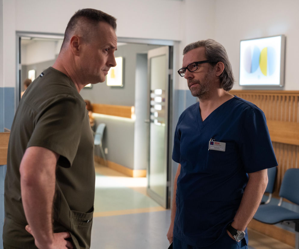 Lekarze Aleś Zagajewski (Paweł Małaszyński) w zielonym ubraniu medycznym i Bart (Piotr Głowacki) w granatowym stroju, rozmawiający w szpitalnym korytarzu. Twarz Zagajewskiego jest wyraźnie zasmucona, Bart patrzy na niego ze spokojem. Scena z serialu Na dobre i na złe, którą można zobaczyć na portalu Super Seriale.