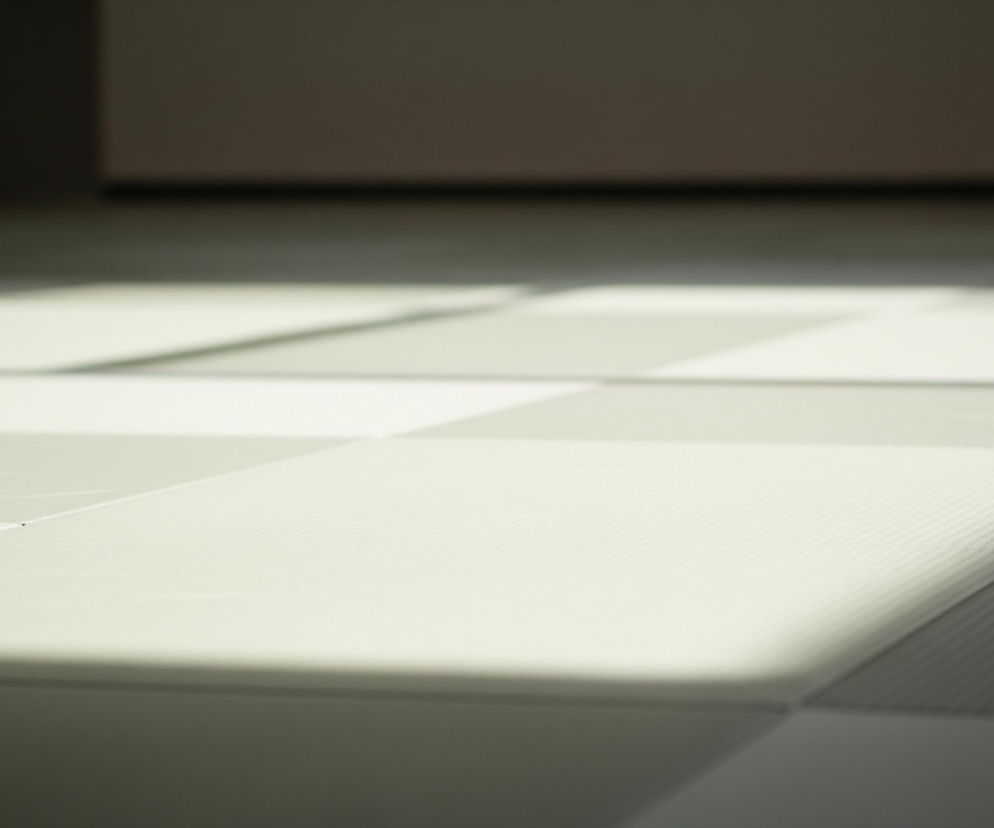 Na pierwszym planie widoczna jest jasna, gładka powierzchnia podłogi, pokryta kwadratowymi panelami w różnych odcieniach szarości i bieli. Panele te tworzą nieregularny wzór, z jaśniejszymi obszarami, które są oświetlone przez ukośne pasy światła. Niektóre panele mają delikatną, prążkowaną teksturę. Tło jest rozmyte i ciemniejsze, ukazując jedynie zarys ścian i podłogi w odległej części pomieszczenia, co sugeruje dużą przestrzeń.