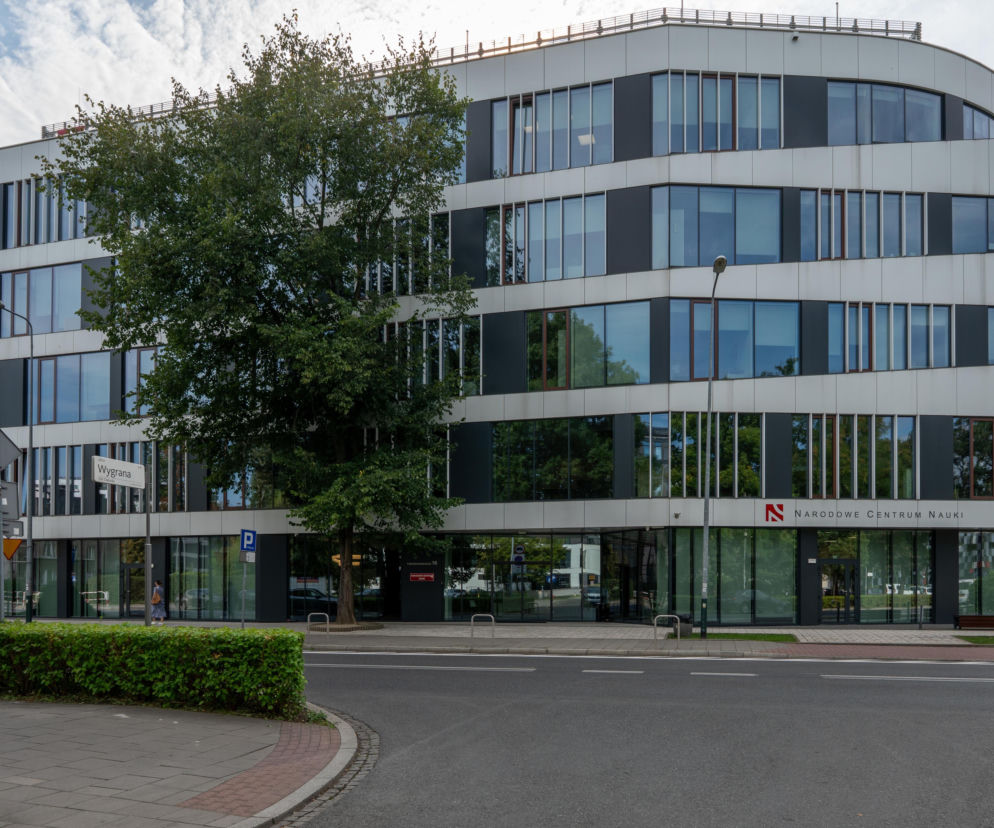 Nowoczesny budynek Narodowego Centrum Nauki w Krakowie, z przeszkloną fasadą i zaokrąglonymi narożnikami, otoczony zielenią. Widoczny znak drogowy Wygrana. Więcej o kontrowersjach wokół NCN przeczytasz na naszym portalu.