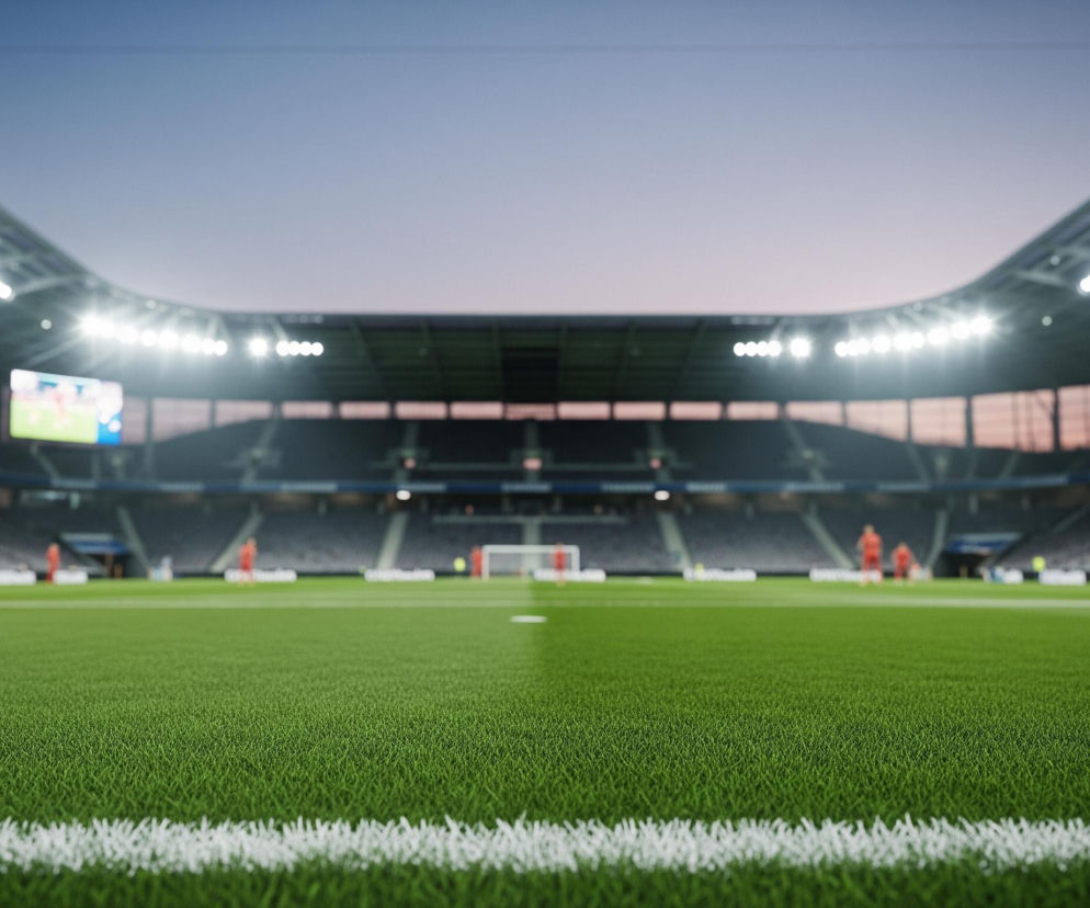 Murawa stadionu piłkarskiego wypełnia pierwszy plan, z wyraźnie widoczną białą linią graniczną boiska na dole kadru. Boisko jest intensywnie zielone i oświetlone jasnym światłem reflektorów zamocowanych na konstrukcji dachu stadionu, które rozchodzą się na boki. W tle widoczne są puste trybuny, zanurzone w cieniu, oraz kilka niewyraźnych postaci w strojach sportowych, częściowo rozmytych. Powyżej trybun rozciąga się wieczorne, lekko zamglone niebo w odcieniach fioletu i błękitu.