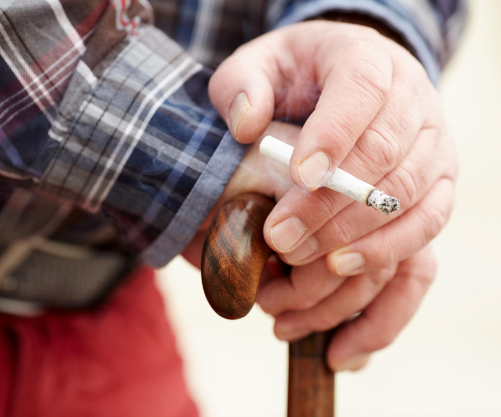 Ręce starszej osoby z siwą skórą i widocznymi zmarszczkami, która trzyma papierosa z palącym się tytoniem i popiołem na jego końcu, opierając się na drewnianej lasce. Obraz symbolizuje palenie a ryzyko choroby Parkinsona, o czym przeczytasz na Poradnik Zdrowie.