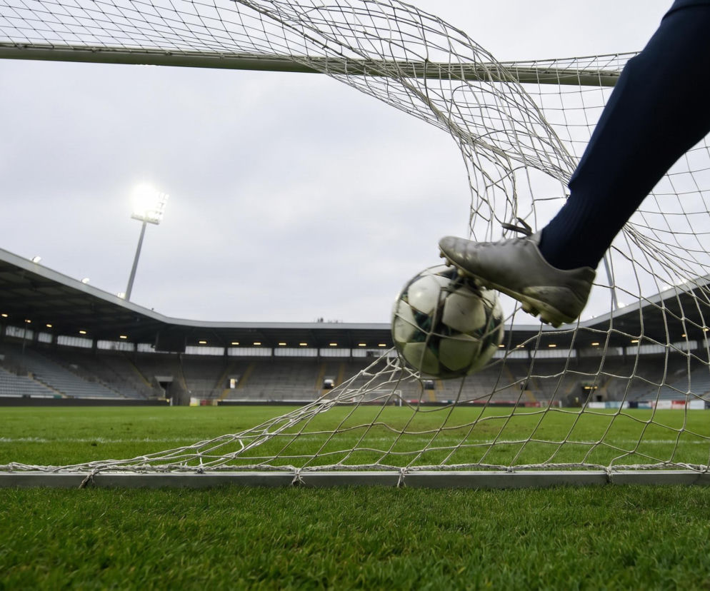 Noga piłkarza w korkach uderza w piłkę, która wpada do bramki, a w tle widać stadion piłkarski ze sztucznym oświetleniem. To ilustracja do artykułu o golach samobójczych, o których więcej przeczytasz na naszym portalu.
