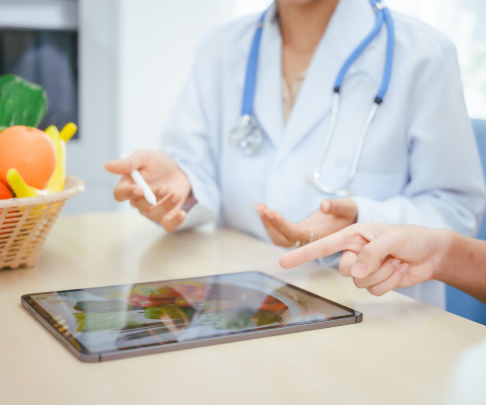 Lekarka z długimi rękawami fartucha i stetoskopem na szyi, rozmawiająca z pacjentką, która wskazuje na tablet z dietą. Obok koszyk pełen owoców i warzyw. Specjalista wyjaśnia, dlaczego po operacji bariatrycznej wraca głód, o czym przeczytasz na Poradnik Zdrowie.