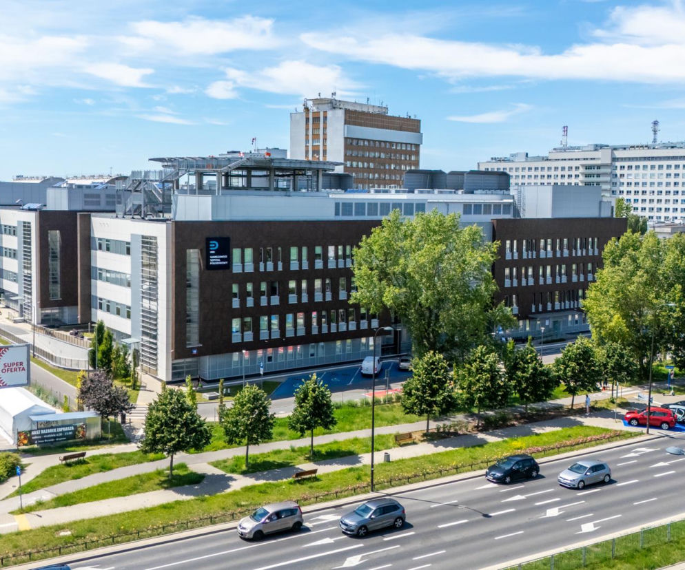 Nowoczesny budynek Warszawskiego Szpitala Południowego widziany z lotu ptaka, z wyraźnym logo placówki na fasadzie. Obok szpitala parking z samochodami, a także mniejsze namioty. To zdjęcie ilustruje temat prokuratorskiego śledztwa w Szpitalu Południowym, o którym piszemy na naszym portalu.
