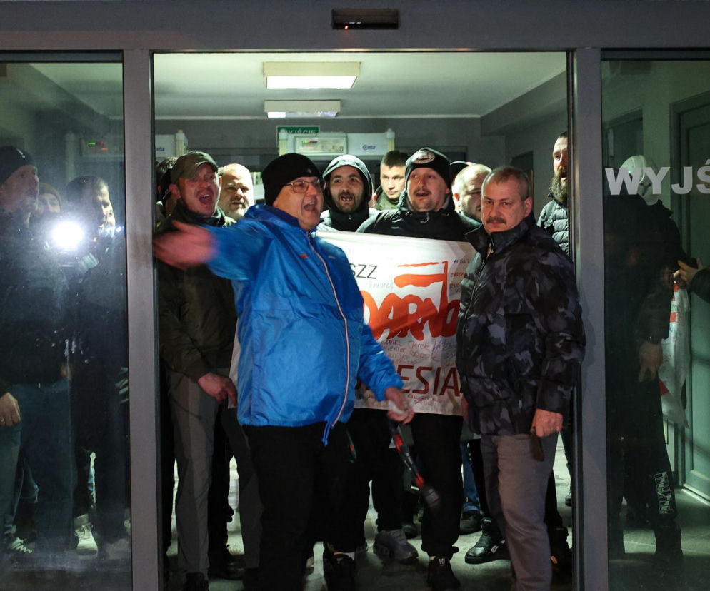 Grupa górników, wychodzących ze świetnie oświetlonego pomieszczenia. Mężczyźni w różnym wieku, niektórzy mają czapki zimowe. W tle widać baner protestacyjny. Wszyscy są oburzeni. Więcej o ich proteście przeczytasz na Super Biznes.