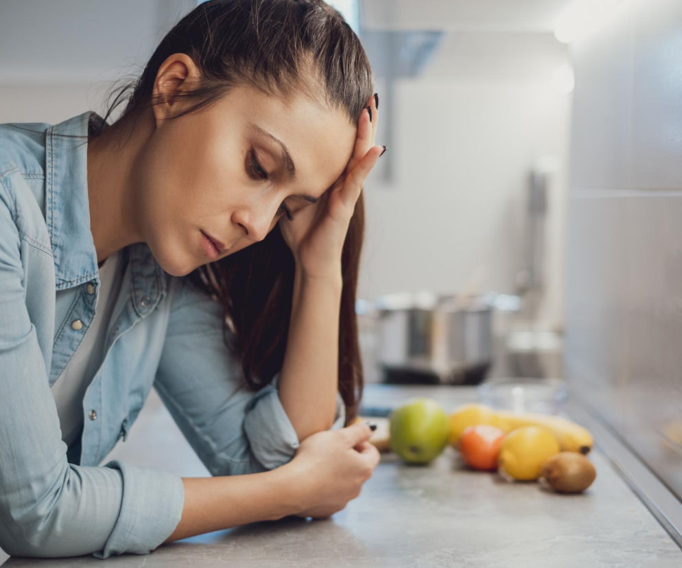 Kobieta zmęczona po posiłku, z opuszczoną głową i dłonią na czole, siedząca przy kuchennym blacie, obok niej owoce. Zdjęcie symbolizuje senność po jedzeniu i poszukiwanie słodyczy, o czym przeczytasz na Poradniku Zdrowie.