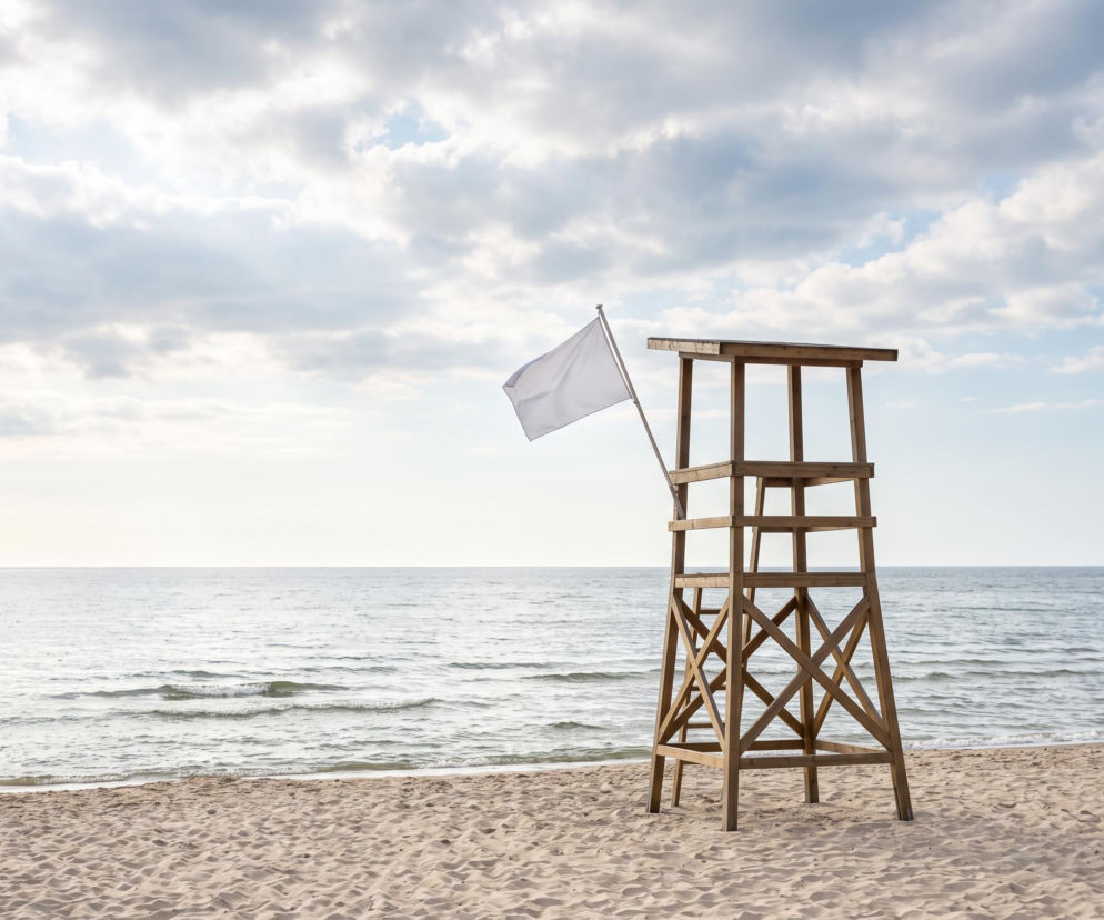 Drewniana wieża ratownicza z białą flagą na pustej plaży. O bezpieczeństwie w Ustroniu Morskim przeczytasz na SE Szczecin.