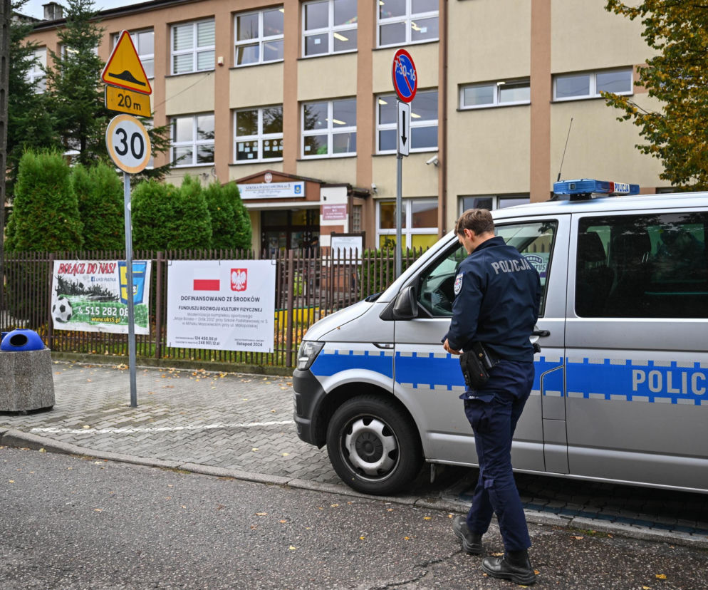 Policjant obok radiowozu przed budynkiem szkoły. O wzmożonych kontrolach i bezpieczeństwie przeczytasz na SE.