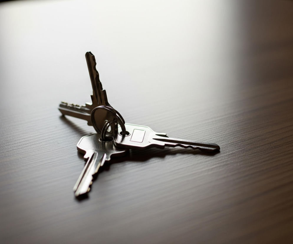 Na brązowym, lekko błyszczącym blacie leży pęk kluczy. Trzy srebrzyste klucze są połączone jednym metalowym kółkiem, z czego dwa leżą płasko, a trzeci, znajdujący się w górnej części kółka, jest lekko uniesiony. Światło pada z lewej, górnej części kadru, rozjaśniając powierzchnię blatu i tworząc cienie rzucane przez klucze.