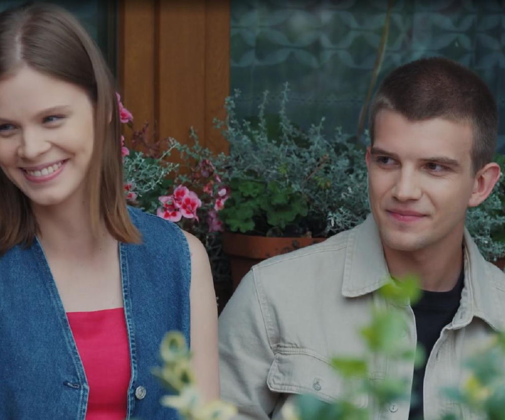 Młoda kobieta o blond włosach, Majka (Matylda Giegżno), uśmiecha się obok młodego mężczyzny, Mateusza (Rafał Kowalski), który patrzy na nią z uśmiechem. Oboje siedzą na zewnątrz, w tle widać rośliny doniczkowe z różowymi kwiatami. Scena z serialu M jak miłość, o której możesz przeczytać więcej na Super Seriale.