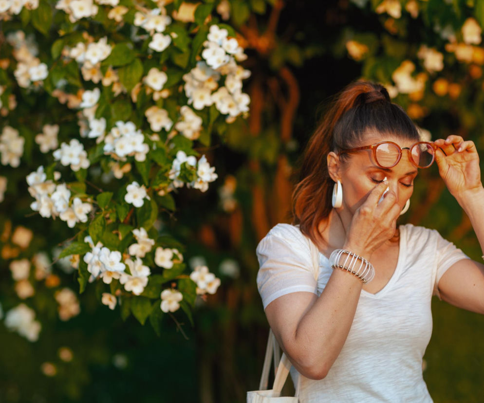 Kobieta w białym T-shircie i okularach na tle kwitnącego jaśminu, ze swędzącymi i łzawiącymi oczami. Alergia na pyłki, takie jak te z krzewów, może powodować katar sienny. Na portalu Poradnik Zdrowie znajdziesz wskazówki, jak ograniczyć kontakt z alergenami.