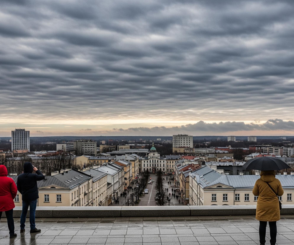 Cztery osoby stojące na płaskim, mokrym, wyłożonym płytkami dachu pod pochmurnym niebem, obserwują panoramę miasta. Dwie osoby po lewej stronie, jedna w czerwonej kurtce z kapturem, druga w czarnej, patrzą w głąb perspektywy ulicy, przy czym ta w czarnej kurtce trzyma telefon komórkowy, robiąc zdjęcie. Dwie osoby po prawej stronie, jedna w musztardowym płaszczu z parasolem nad głową i torbą przewieszoną przez ramię, oraz druga w czarnej kurtce z otwartym czarnym parasolem, również patrzą w tę samą stronę. Poniżej rozciąga się miejski krajobraz z długą, prostą ulicą, wzdłuż której stoją budynki o jasnych elewacjach i ciemnych dachach. W oddali widoczne są wyższe budynki mieszkalne, drzewa bez liści oraz szare, niskie chmury pokrywające większość nieba, z jaśniejszym pasmem u horyzontu.