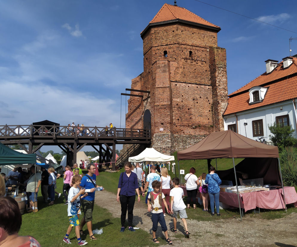 Uczestnicy V Festiwalu „W Zamkowej Kuchni na Szlaku Książąt Mazowieckich” w Liwie, spacerujący po dziedzińcu przy historycznej wieży z cegły z czerwoną dachówką oraz białym budynku z czerwonym dachem. Na pierwszym planie stoiska festiwalowe. Więcej o wydarzeniu na naszym portalu.
